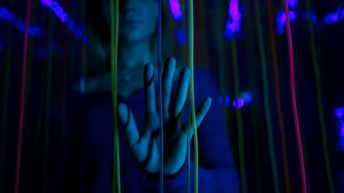 A young woman interacts with colorful neon ropes in a dark, artistic setting, highlighting her hand in close-up.