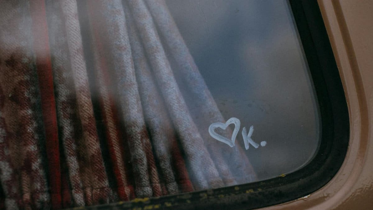 A close-up of a van window with a heart symbol and initial 'K' on a rainy day.