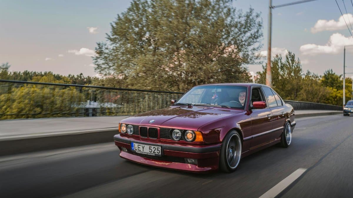 A red BMW E34 sedan driving on a scenic road with trees and clear sky in the background.
