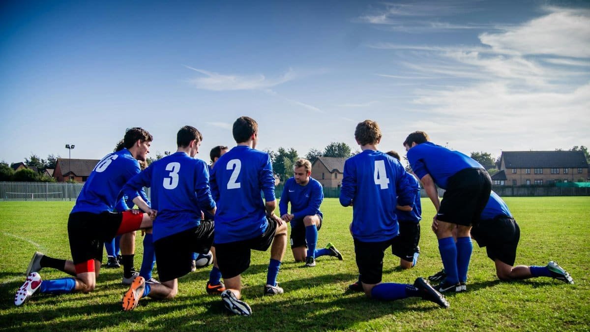 A group of male football players in blue uniforms huddle on a grassy field before a match.