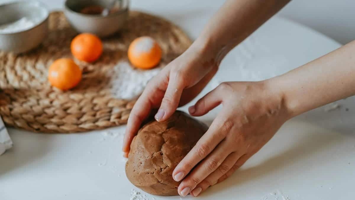 Hands kneading brown dough on a white table, surrounded by oranges and kitchen utensils.
