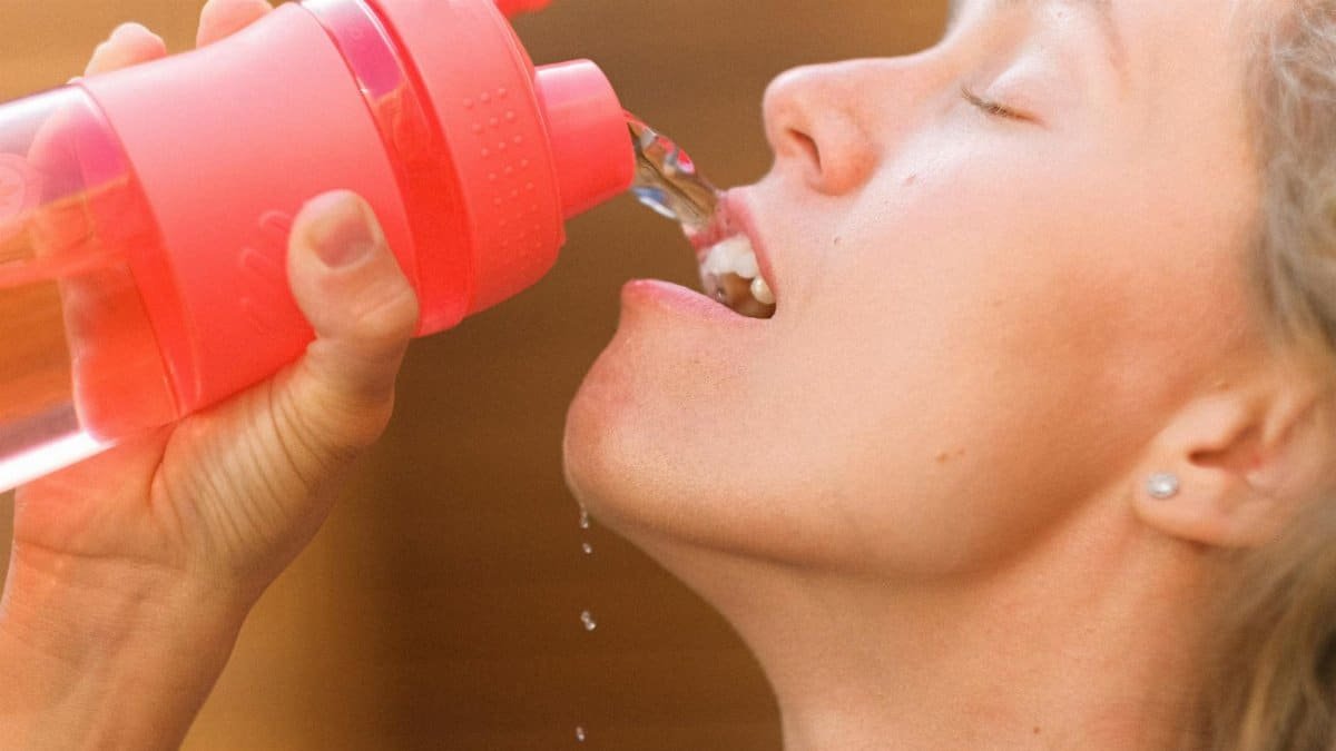 Crop tired young female pouring refreshing clean water into mouth from reusable bottle after intense training