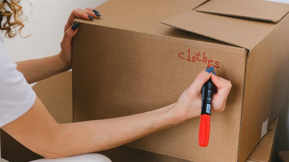 Close-up of a woman labeling a cardboard box with a red marker indoors.