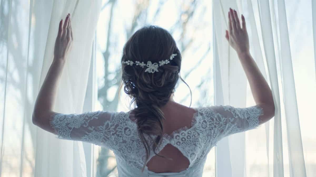 Beautiful bride in lace dress looks out a sunlit window, capturing a moment of serene elegance.