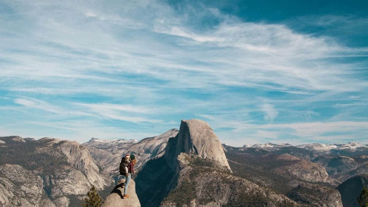 A woman and child stand atop Half Dome, Yosemite, with breathtaking views of the mountain range.