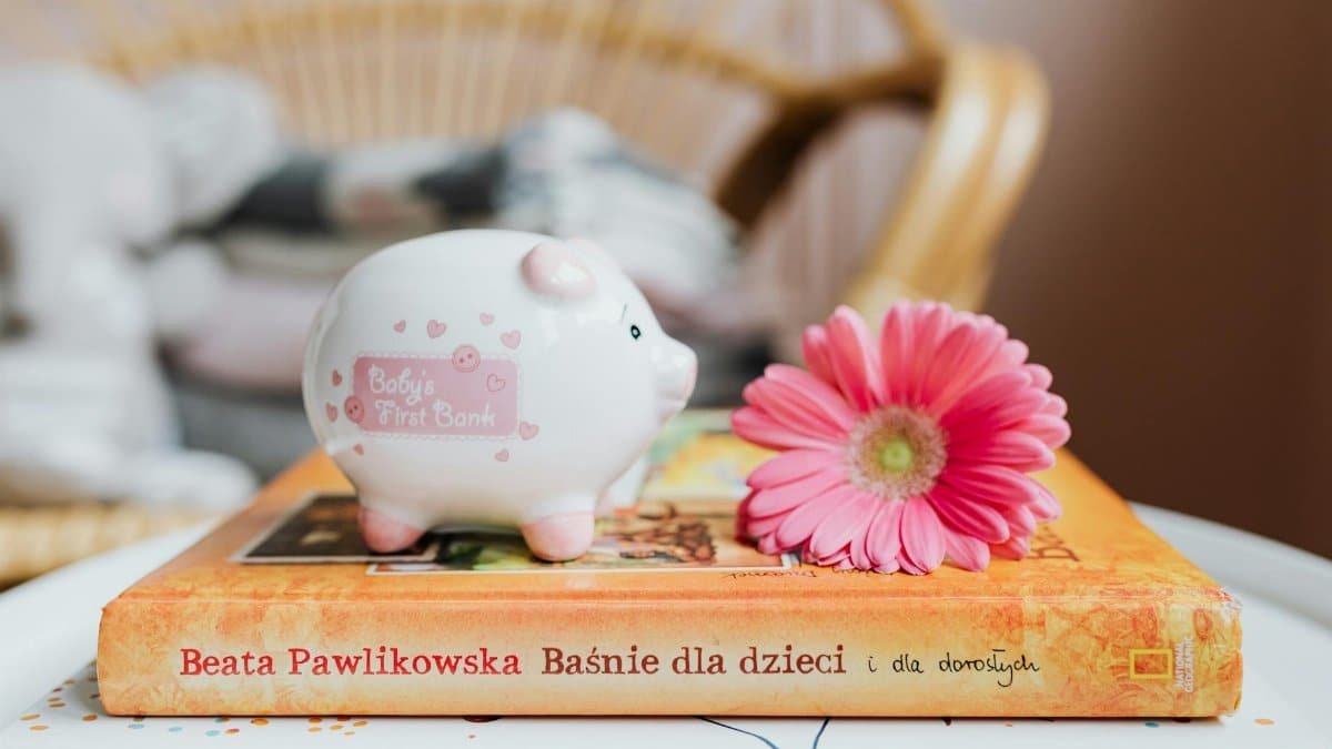 A piggy bank and pink gerbera flower rest on colorful children's books, suggesting childhood savings.