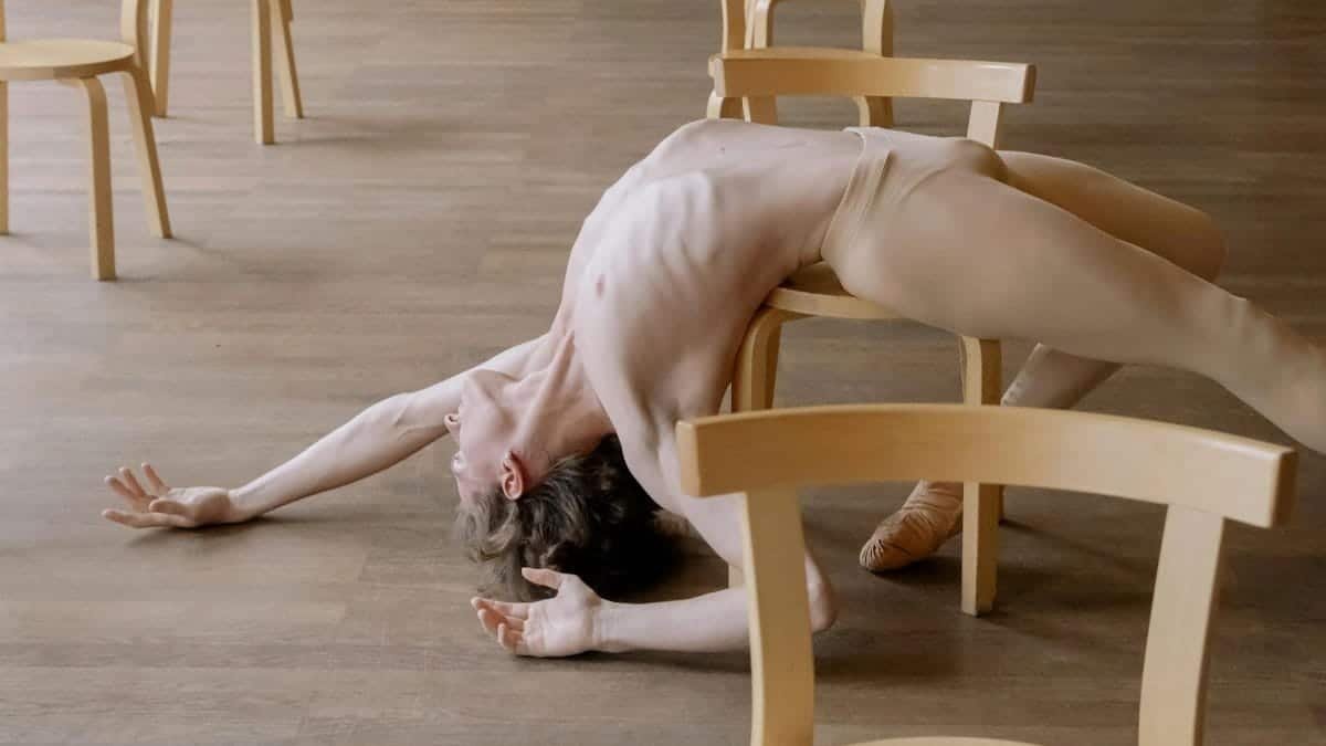 Male dancer in a modern pose on wooden floor with chairs around, showcasing flexibility.