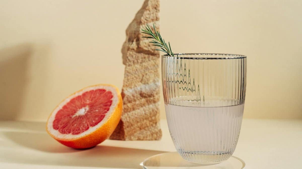 A clear glass with a grapefruit beverage and rosemary garnish on a minimalist beige backdrop.