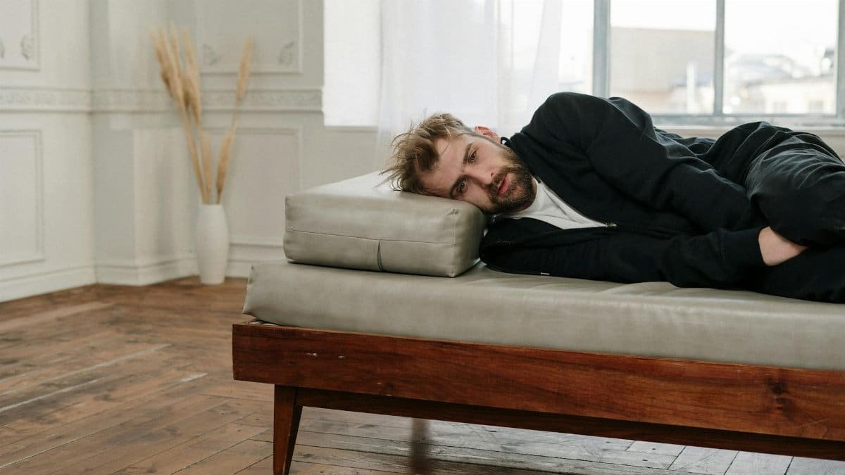A bearded man lying on a couch in a bright, minimalistic room, looking contemplative.