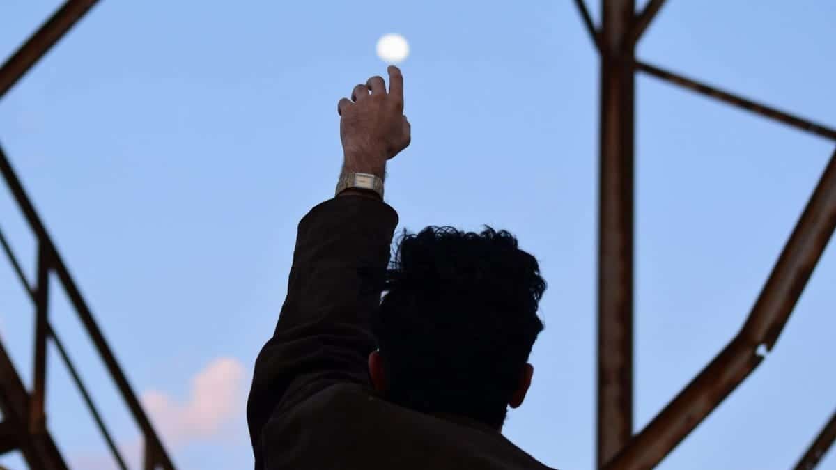 A man silhouetted against the morning sky reaches towards the moon with an outstretched hand.