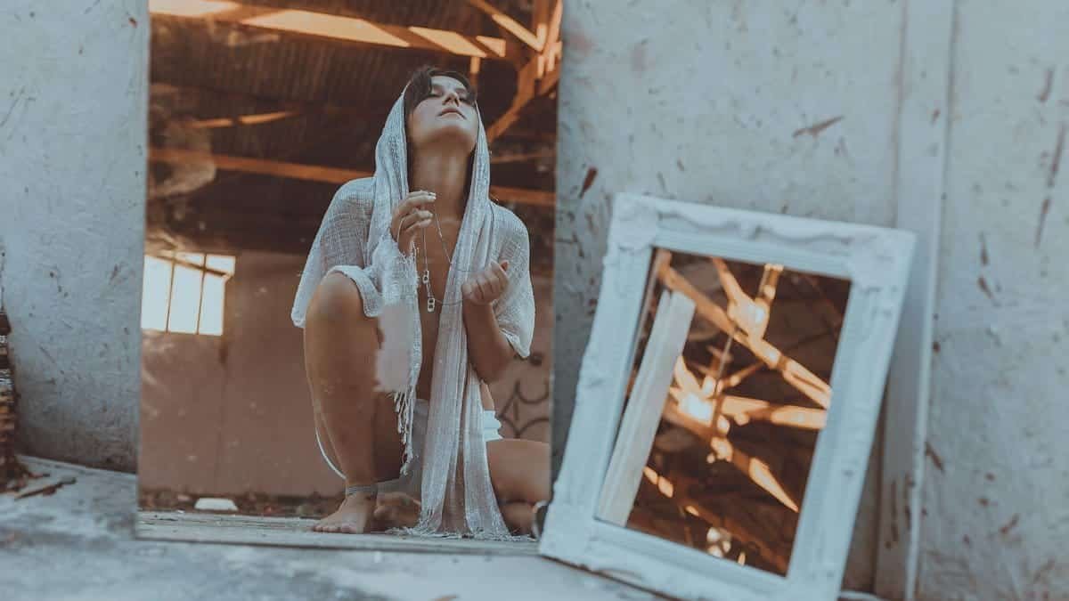 A woman in a hooded garment crouches in front of a mirror in an abandoned building, contemplating.
