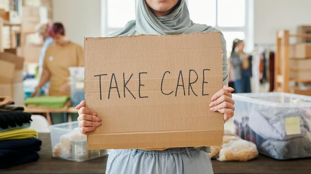Smiling volunteer in hijab holding a 'Take Care' sign at a charity event indoors.