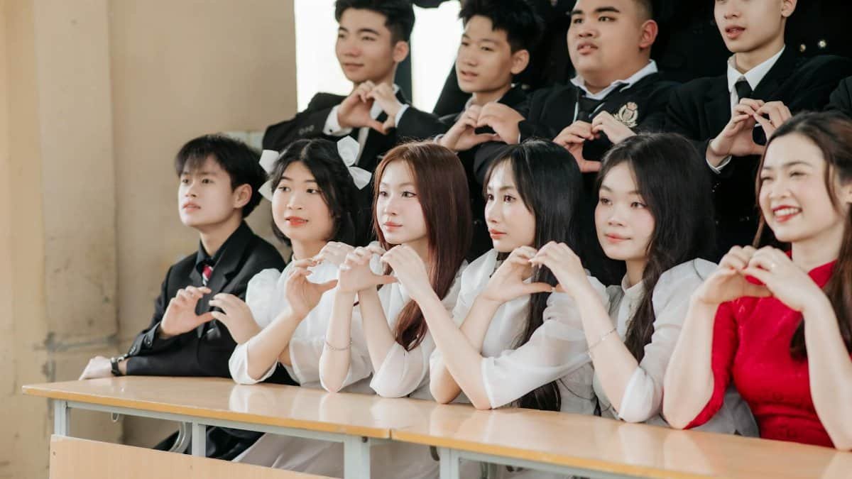 Students sitting at desks forming heart shapes with hands in a classroom setting, wearing formal attire.