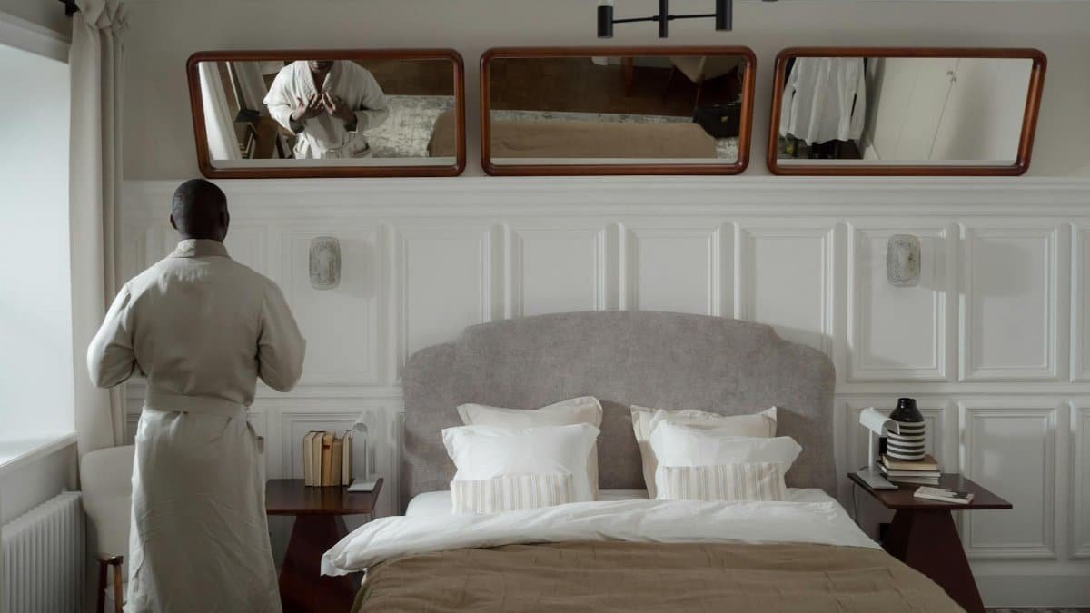 Adult man in bathrobe adjusting clothing in a stylish bedroom setting with mirrors.