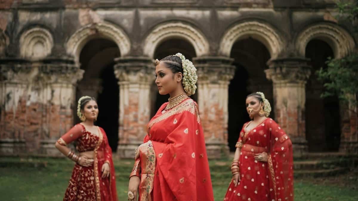 Three Indian women in vibrant red sarees pose gracefully near ancient temple architecture.