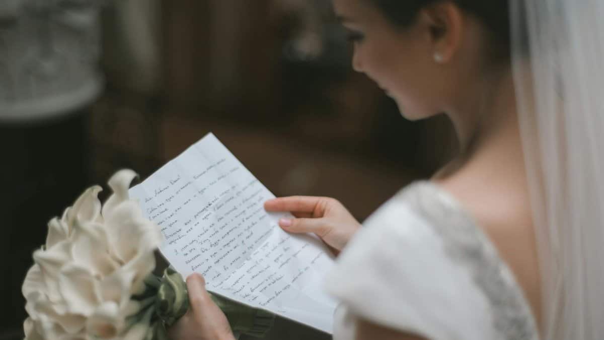 Back view of crop charming bride in white dress and veil reading wedding vow on paper sheet with bouquet of arum lilies during weeding ceremony