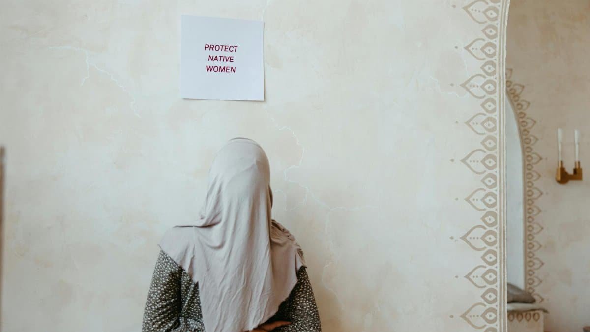 A woman in a hijab stands facing a wall with a sign reading 'Protect Native Women'.