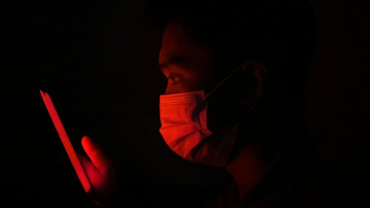 A person wearing a face mask uses a smartphone illuminated by red light in a dark setting.