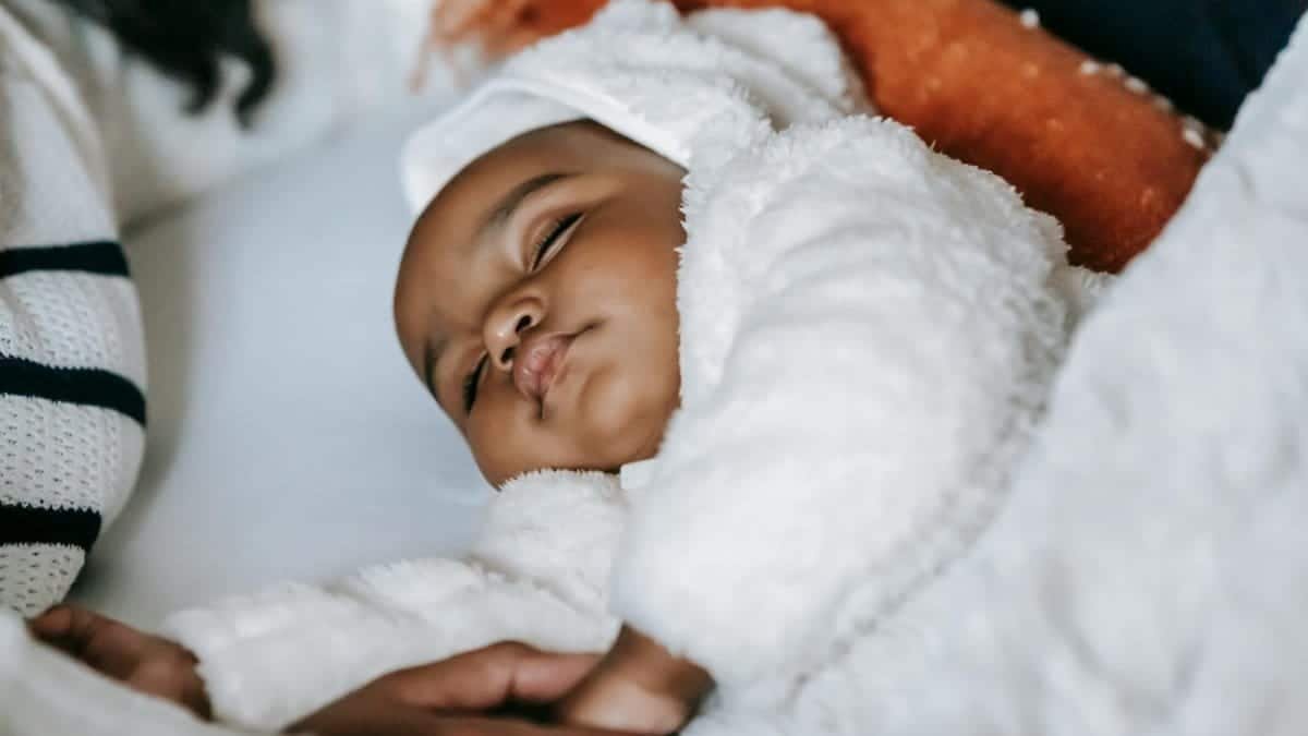 From above of crop anonymous African American mother holding hand of cute newborn baby in warm clothes sleeping on cozy bed