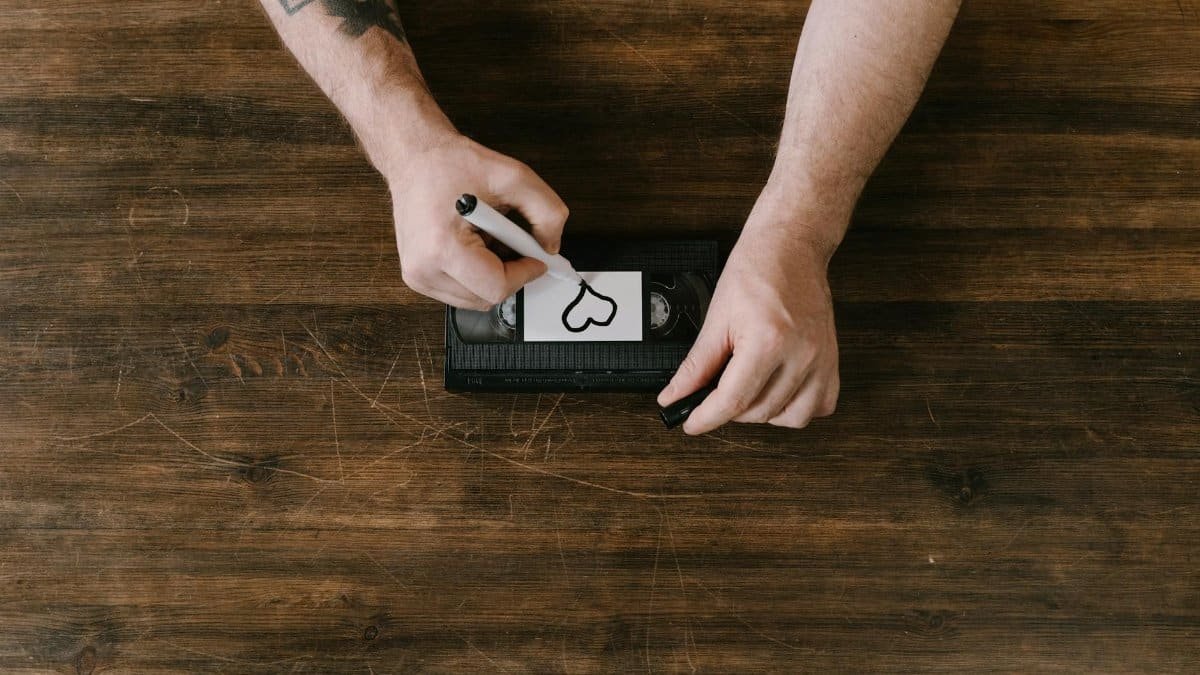 Close-up of hands drawing a heart on a VHS tape label with a marker on a wooden surface.