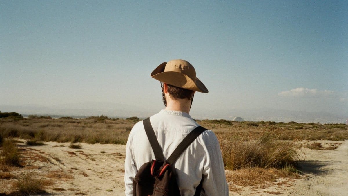 A traveler with a backpack and hat explores a wide desert landscape under a clear blue sky.