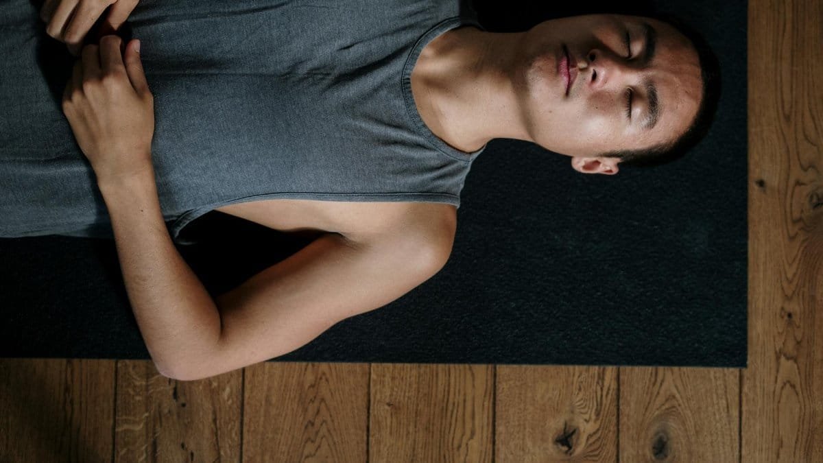 A man lies on his back in a yoga studio, relaxing on a mat in natural light.