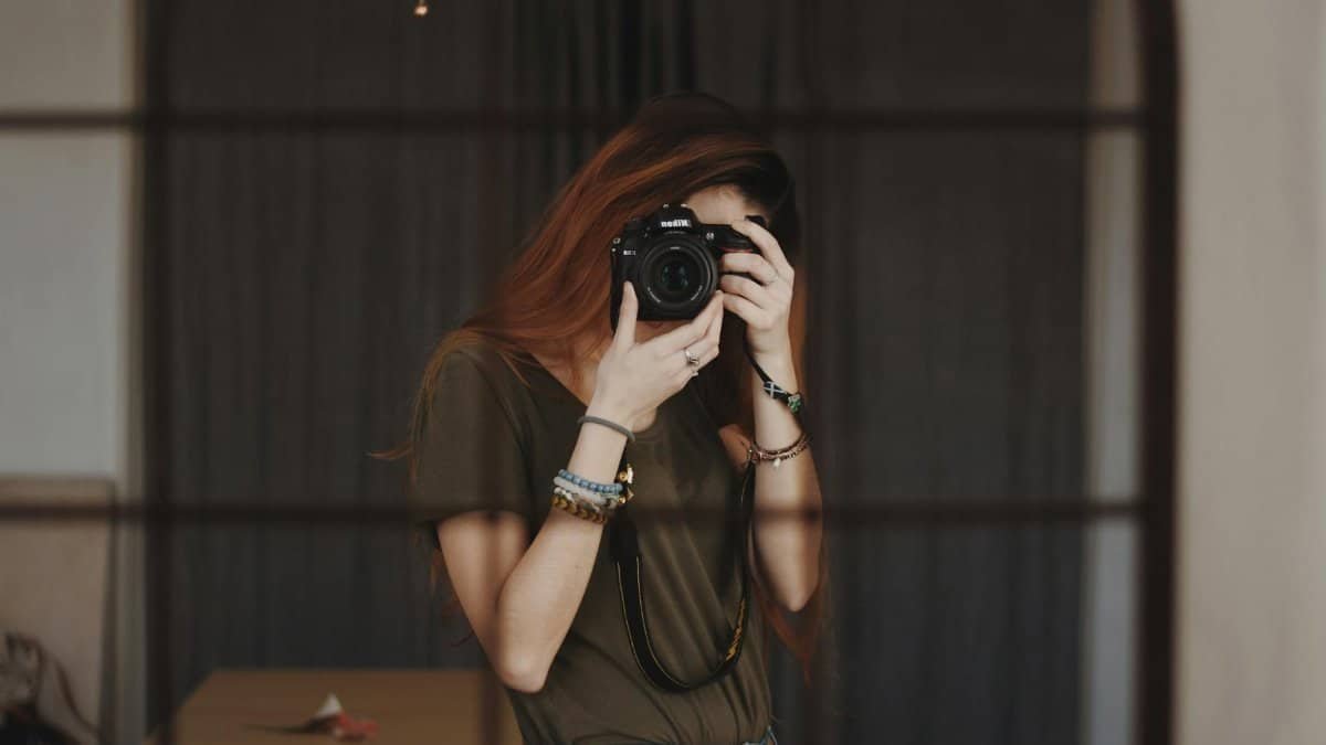 A female photographer capturing a self-portrait with a DSLR camera in an indoor setting.