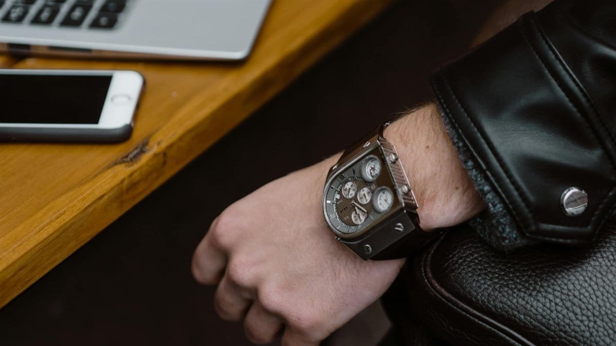 Close-up of a stylish metal wristwatch on a person's wrist with a leather jacket, next to a smartphone and laptop.