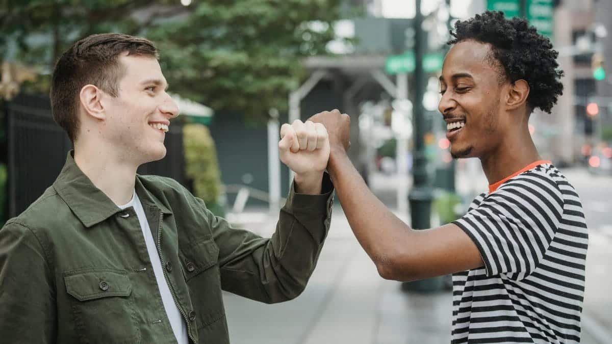Side view of joyful young multiracial male friends in casual outfits smiling while bumping arms after meting on city street