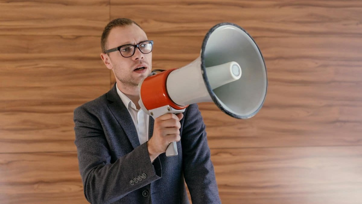 Man in suit holding a megaphone, making a public announcement indoors.