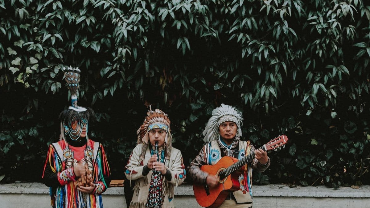 Three men in Native American attire playing instruments outdoors, showcasing cultural traditions.