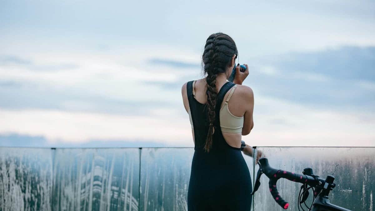 Back view of unrecognizable sportswoman drinking beverage from mug near bicycle and fence while having break