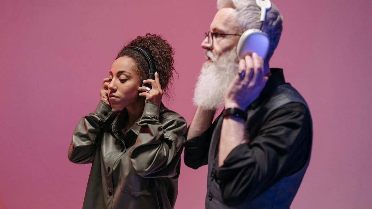 Man and woman listening to music on headphones against a pink background.