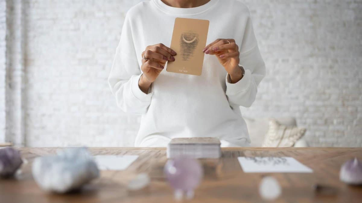 A person wearing white holds a tarot card with gemstones on a wooden table for divination.