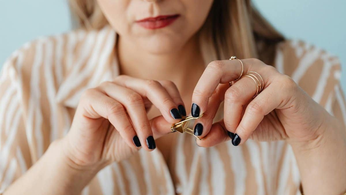 Close-up of a woman's hands fidgeting with a gold paperclip, showcasing painted nails and jewelry.