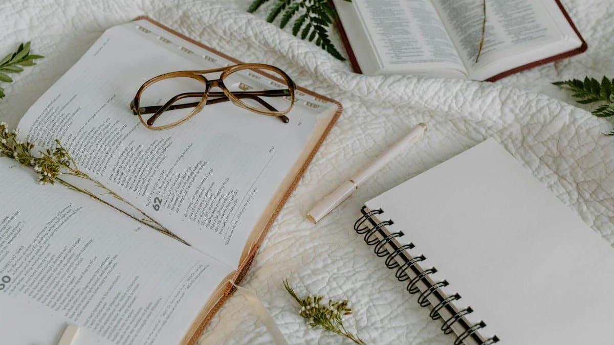 Open Bible with glasses, notepad, and pen on a white quilt, evoking study and reflection.