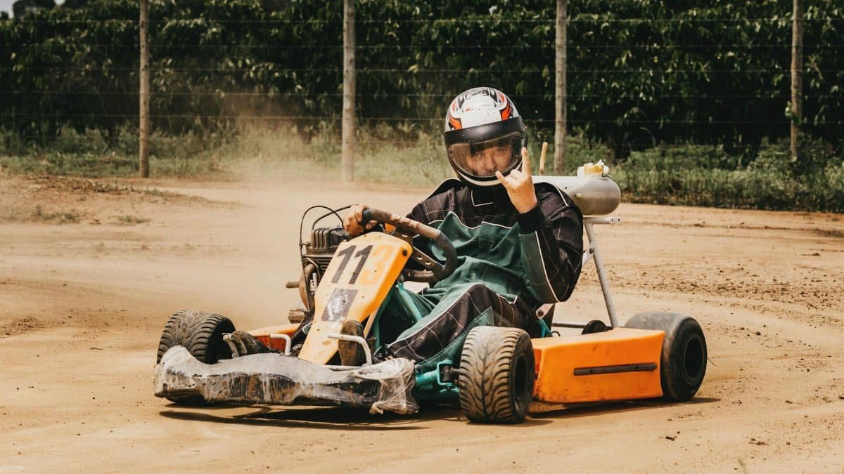 A driver in protective gear racing a go-kart on a sandy outdoor track with a fence backdrop.