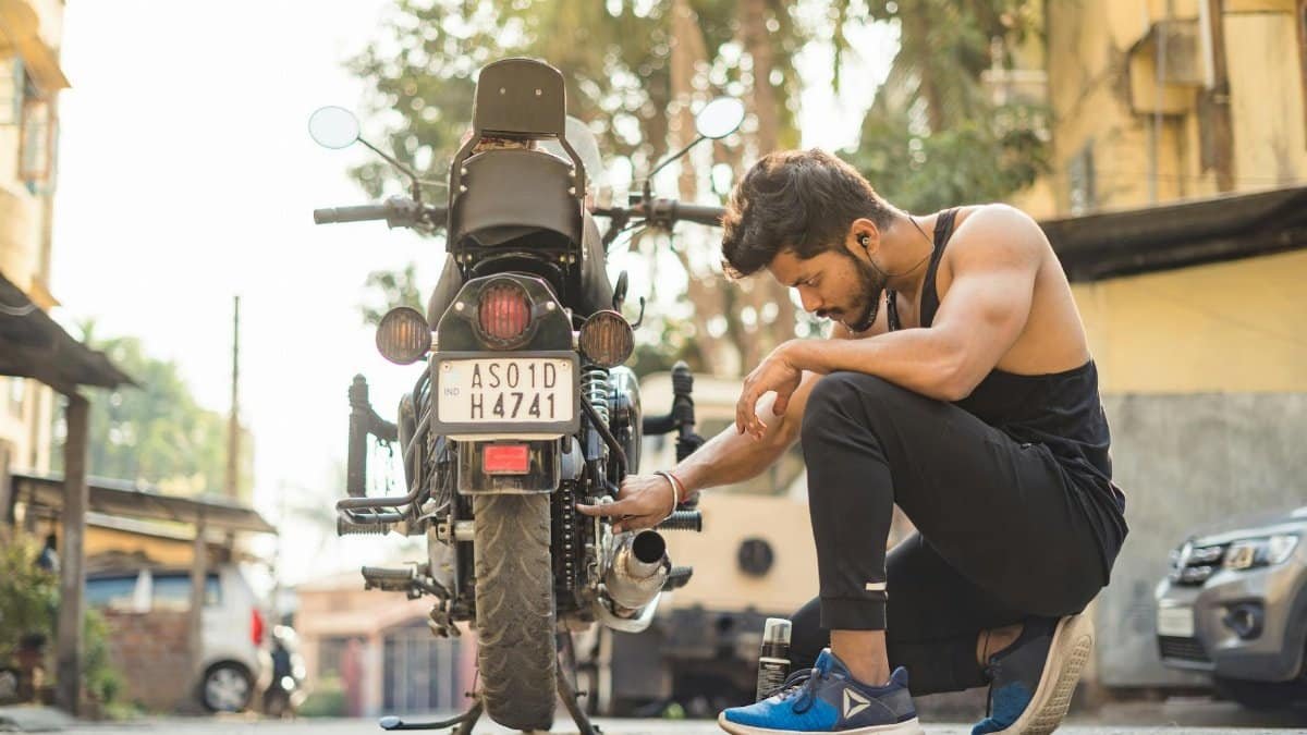 A muscular man in black attire fixing a motorcycle on a sunlit street, showcasing focus and concentration.