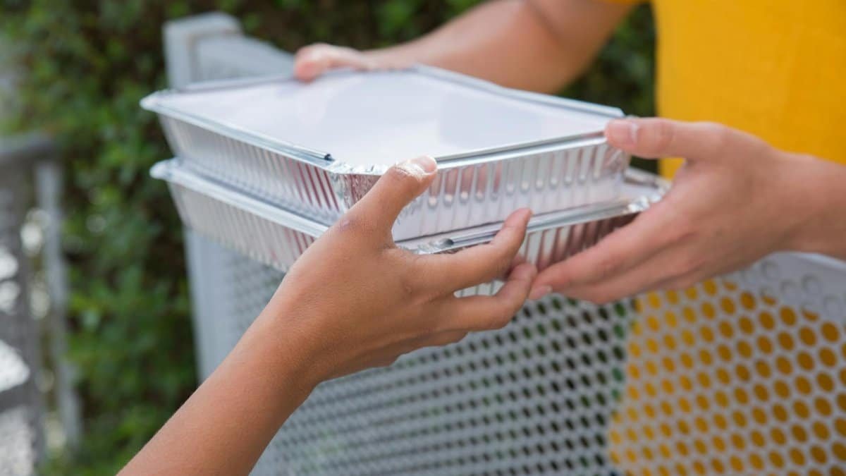 Close-up of two people exchanging aluminum food containers over a metallic fence outdoors.