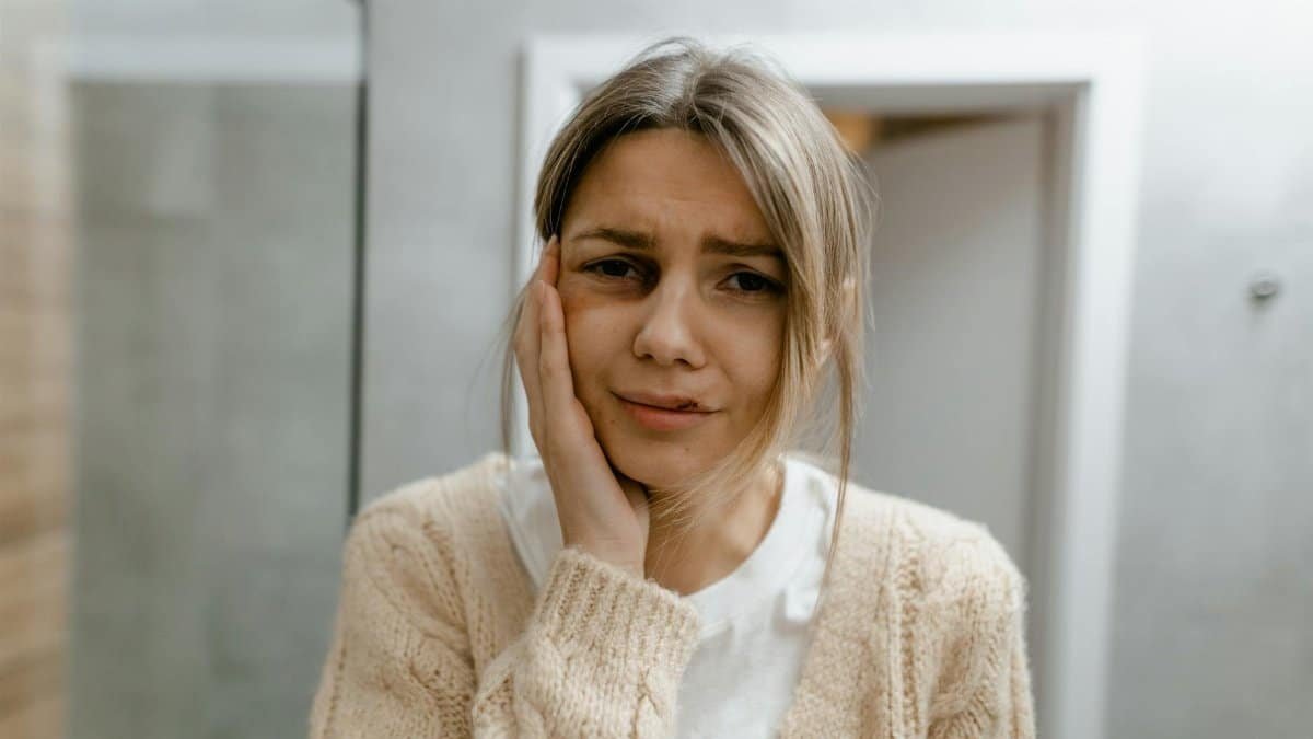 A worried woman stands indoors, touching her face, displaying signs of discomfort.
