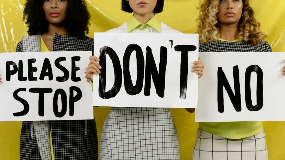 Three diverse women holding protest signs against yellow background promoting unity and activism.