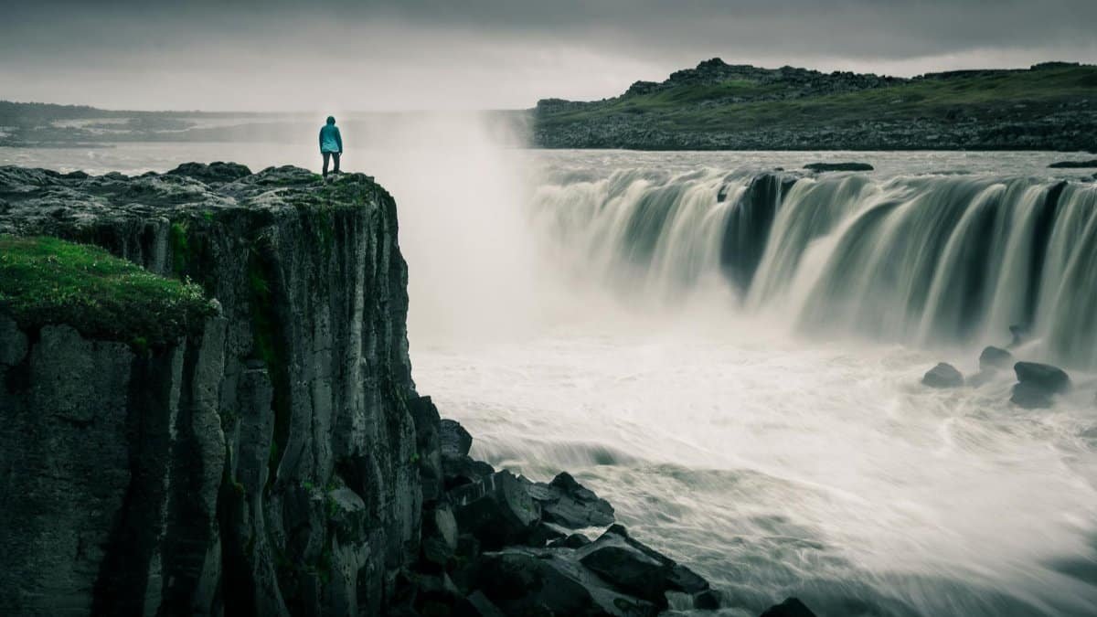 Lone adventurer standing atop cliffs beside a powerful waterfall, showcasing nature's grandeur.