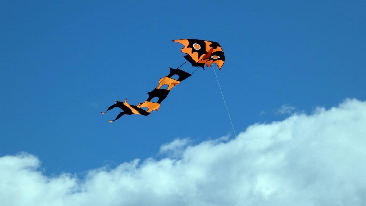 Bright orange and black kite flying high against a clear blue sky with clouds.