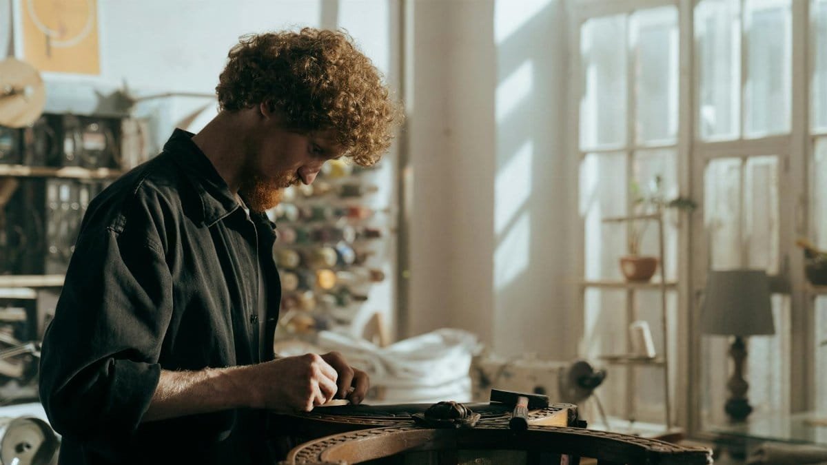 A redheaded craftsman focuses on restoration work in a sunlit workshop, surrounded by tools and materials.