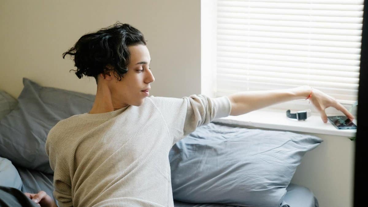 Young ethnic male adolescent taking mobile phone from windowsill after awakening in morning in bedroom
