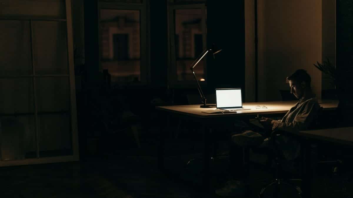 A man working late at night in a dimly lit office, with a bright laptop screen.