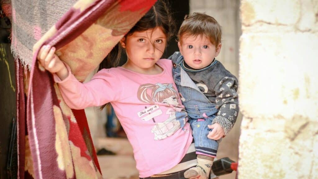 A young girl holding a baby stands in a doorway in Idlib, Syria, capturing resilience and innocence amidst conflict.