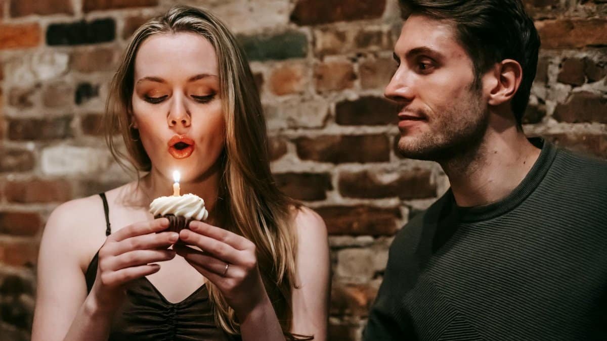 Young couple in elegant outfits in restaurant while blowing in candle on small cupcake and celebrating birthday near brick wall