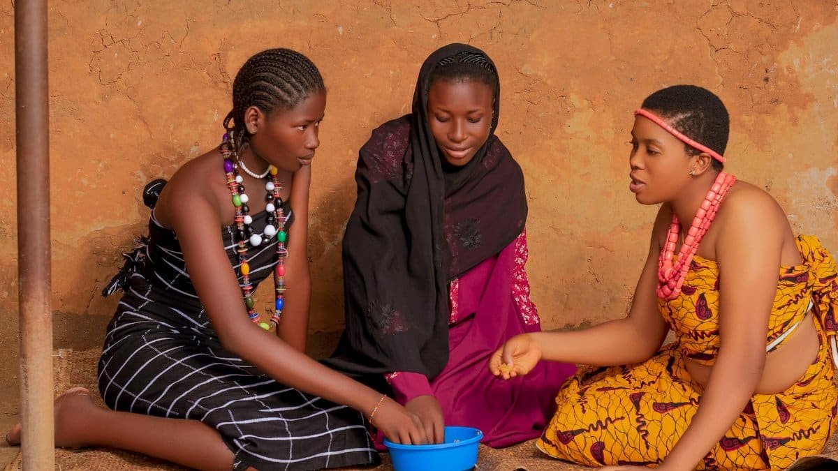 Three African women in traditional attire sit together, engaged in conversation.