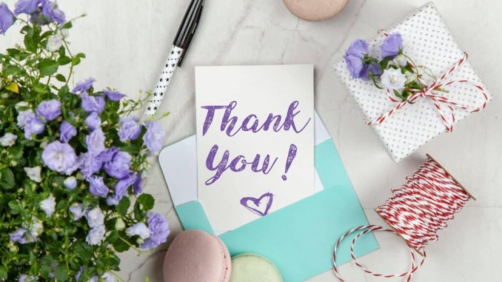 A beautifully arranged thank you card with lavender flowers, macarons, and gift package on a marble desk.